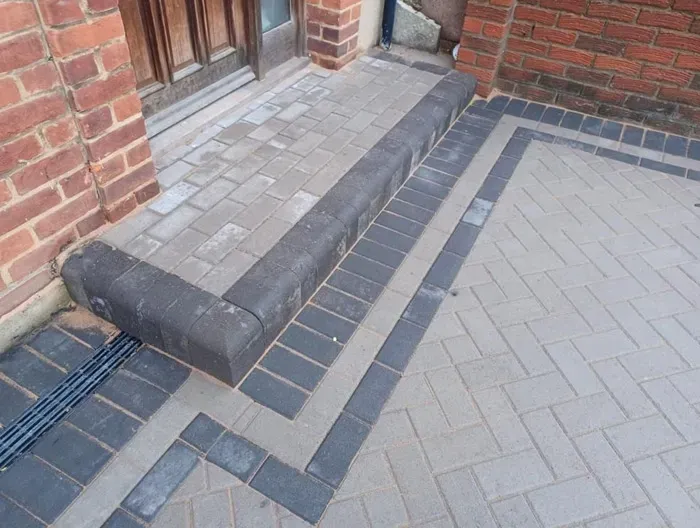 Close-up of a new block paved driveway entrance with light grey blocks, dark grey border, matching kerb stone, and a linear drainage grate.