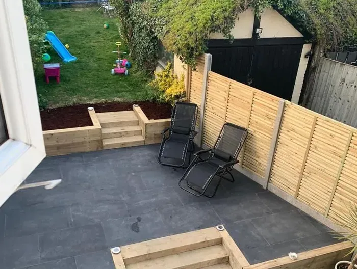 Newly landscaped garden with dark grey patio, wooden steps, raised garden bed, green lawn, and modern wooden fencing.