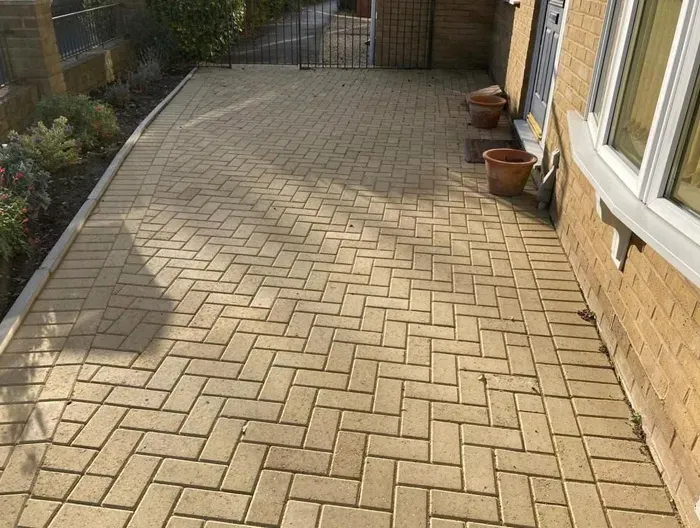 A well-maintained, light-coloured block-paved driveway in a herringbone pattern, bordered by a subtle kerb and a garden bed, leading to the entrance of a brick house.