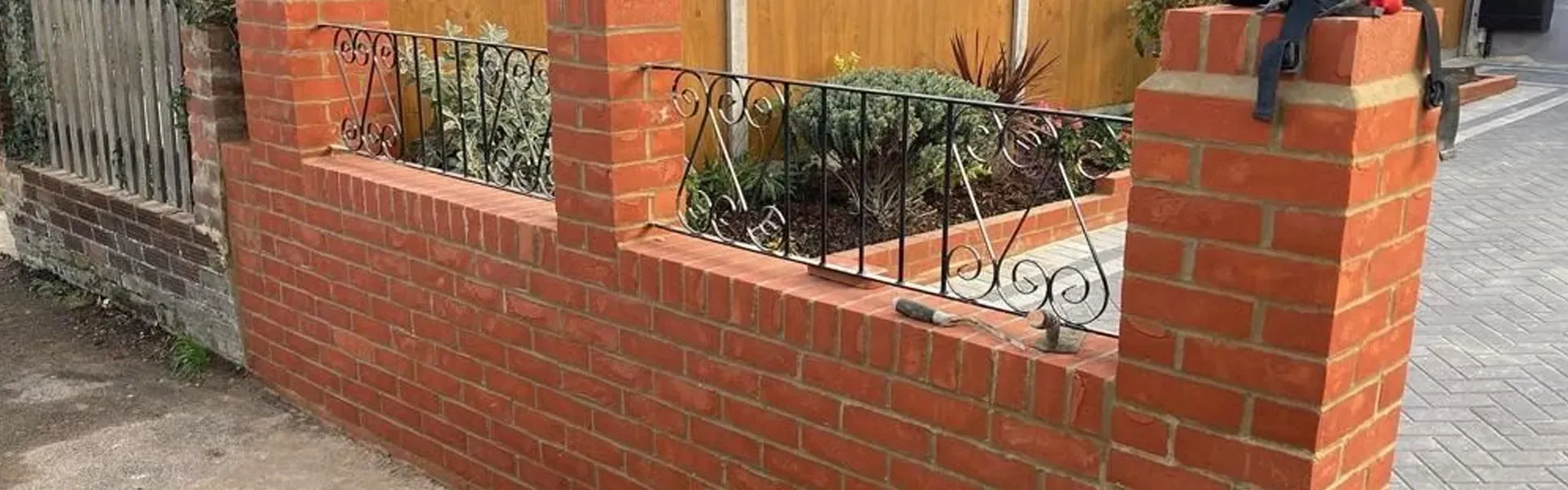 A freshly built red brick garden wall with elegant black wrought iron railings and a visible section of a grey block-paved driveway.