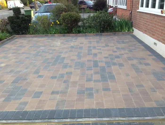 A newly laid block paved driveway in alternating brown and grey blocks with a dark grey border, leading up to a house and garden.