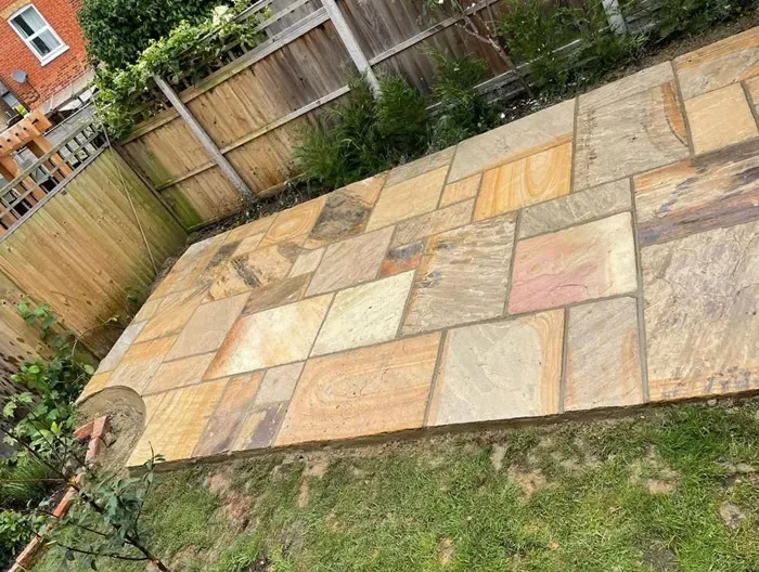 A beautiful, newly laid Indian Sandstone patio in a garden, showcasing varied coloured natural stone paving slabs next to a green lawn and a timber fence.