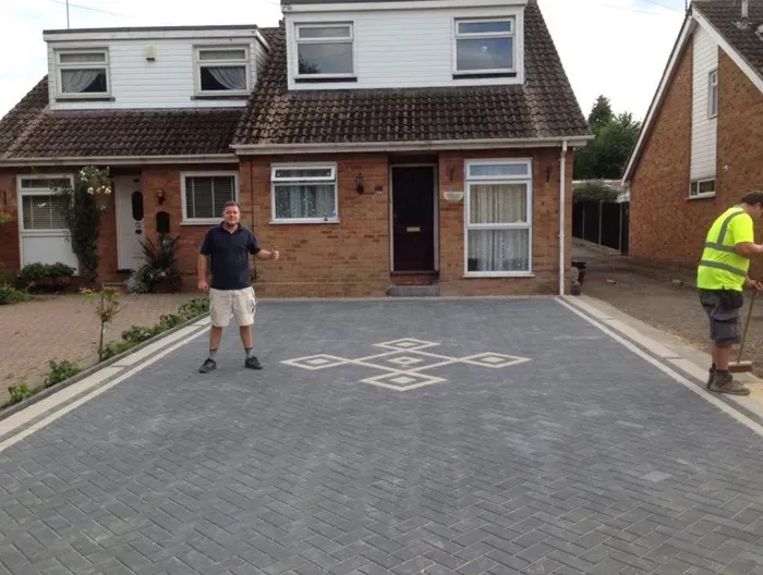 Newly installed grey block paving driveway with a decorative pattern in front of a brick house, with a man giving a thumbs-up and another person sweeping.