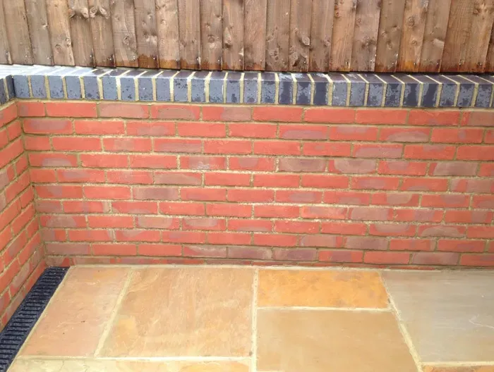 New red brick garden wall with dark coping above a natural stone patio and a linear drain.