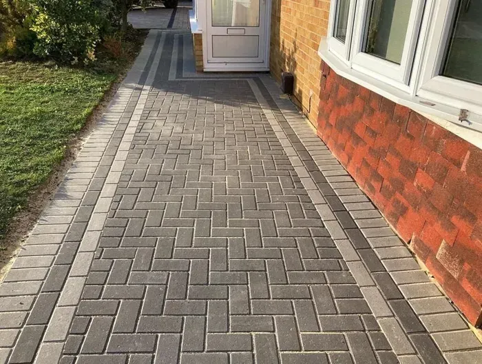 Newly laid block paving path or driveway in grey and lighter grey herringbone pattern with a contrasting border, leading to a house with a white front door and bay window.