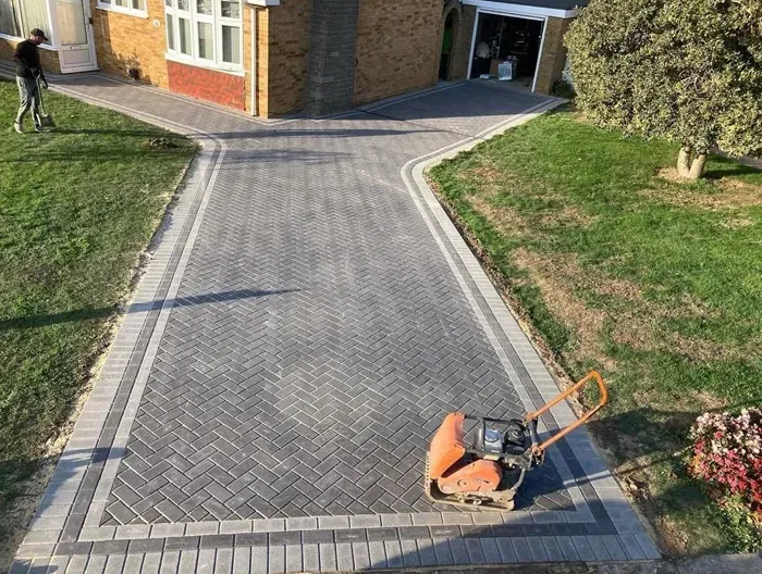 Newly laid block paving driveway with a dark grey herringbone pattern and lighter grey border, with a compaction plate on the surface, next to a green lawn and house.